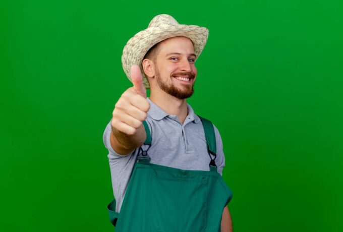 smiling young handsome slavic gardener in uniform and hat looking at camera showing thumb up isolated on green background with copy space
